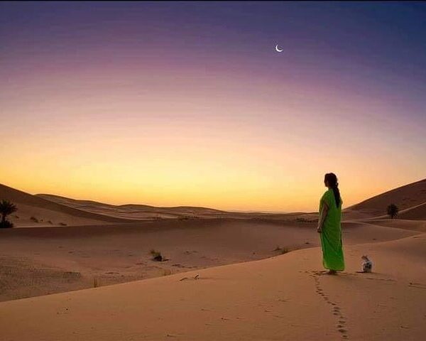 Traveler standing on golden Sahara Desert dunes at sunset with crescent moon in the sky.