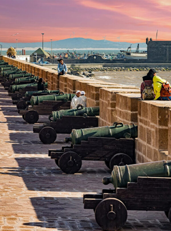 Row of old cannons along a seaside stone wall with people walking and ocean views at sunset.